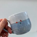 A hand is holding up a blue and white mug in front of a white background. The mug has a small swimmer on the side - what is visible is their red hat, orange tow float and a blue swimsuit