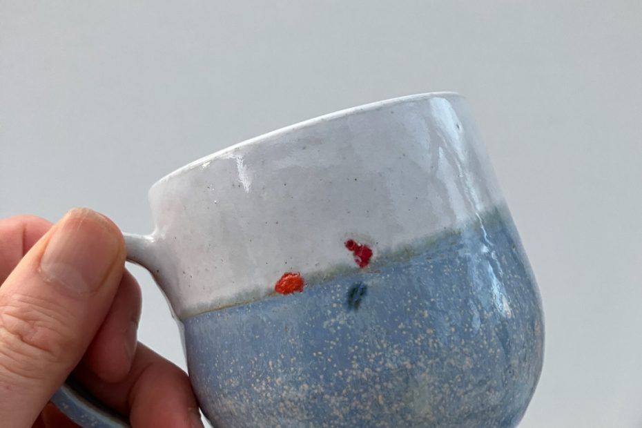 A hand is holding up a blue and white mug in front of a white background. The mug has a small swimmer on the side - what is visible is their red hat, orange tow float and a blue swimsuit