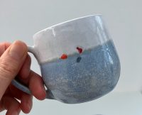 A hand is holding up a blue and white mug in front of a white background. The mug has a small swimmer on the side - what is visible is their red hat, orange tow float and a blue swimsuit
