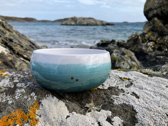 A sea-green and white curved bowl is balanced on the rocks with the sea behind