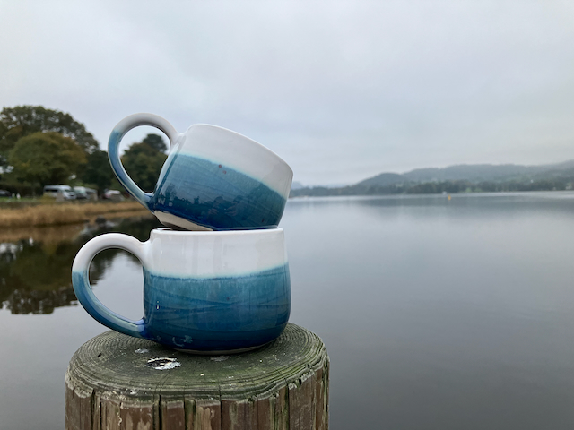 Two mugs in dark blue and white glaze are balanced on top of a wooden post with Ullswater behind
